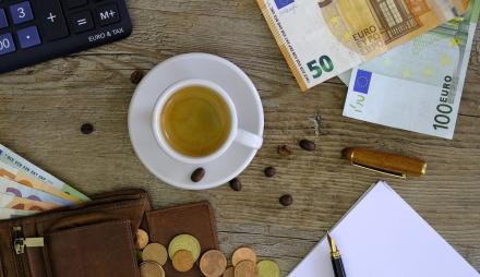 Gefüllte Kaffetasse auf einem Tisch, auf dem Geldscheine, Münzen und ein Notizblock mit Stift liegen