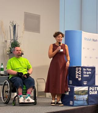 Dr. Tina Denninger mit einem Mann im Rollstuhl auf der Bühne der IHK Potsdam beim Inklusiven Kaffeeklatsch 2026