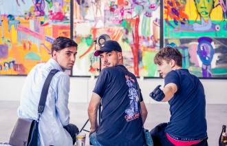 3 junge Männer sitzen auf einer Bank vor einer bunt bemalten Wand