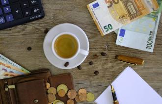 Gefüllte Kaffetasse auf einem Tisch, auf dem Geldscheine, Münzen und ein Notizblock mit Stift liegen
