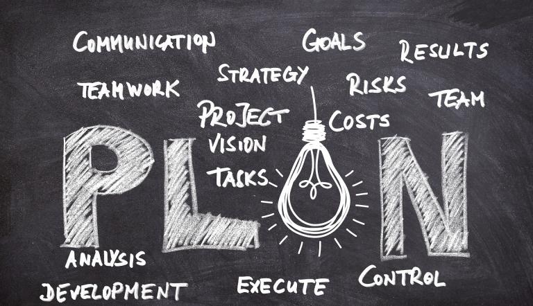 Tafel, auf der mit weißer Kreide "Plan" in großer Schrift steht sowie weitere Bestandteile des Businessplans