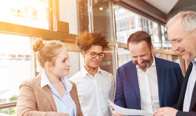 4 Personen im Büro, die freundlich lächeln und auf einem Blatt Papier schauen